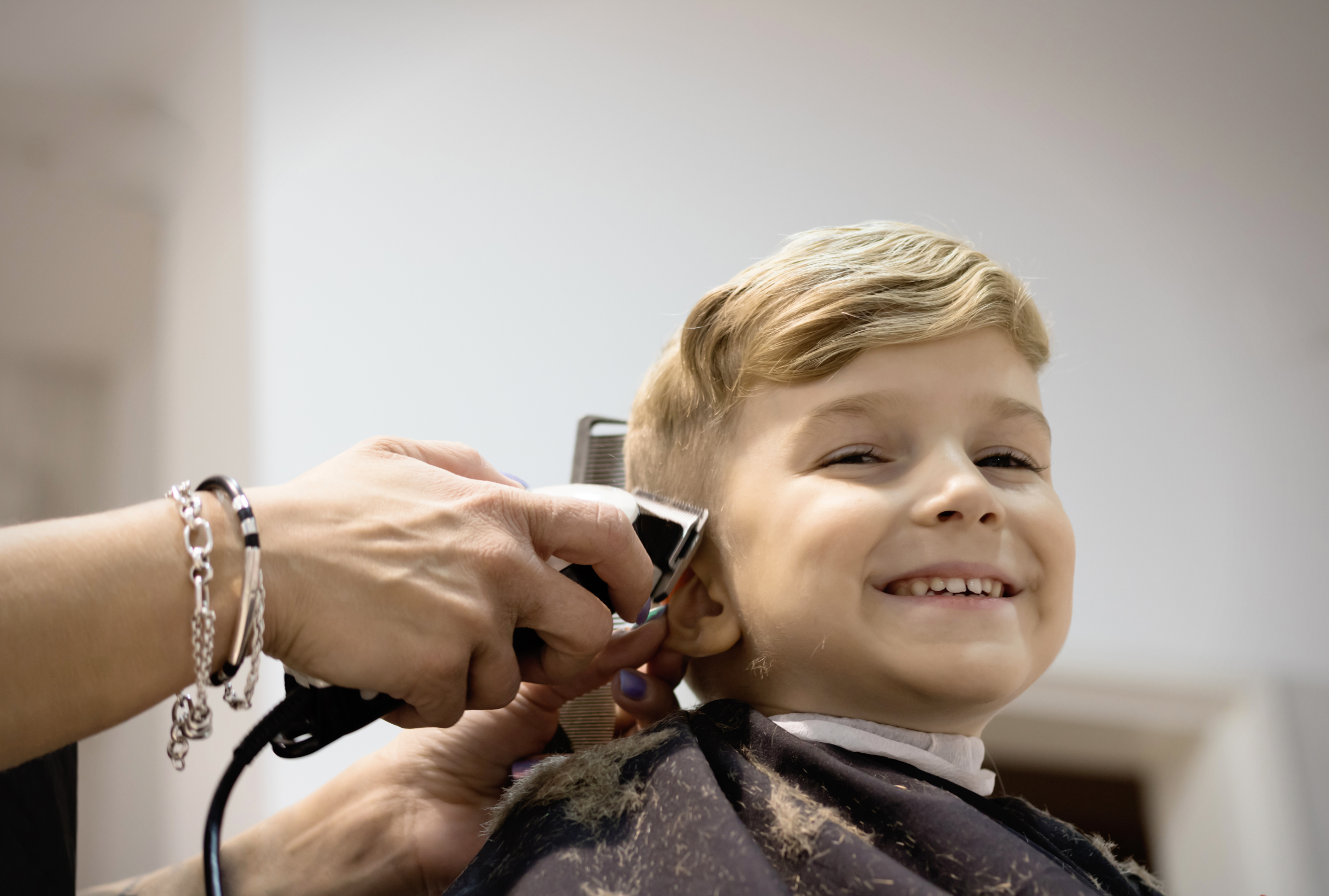 A smiling young boy sits in a salon chair wearing a cape while a stylist trims his hair with clippers, representing strategies that help a child with autism handle haircuts.