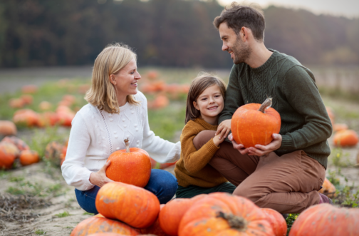 A smiling mother, father, and young child kneel together in a pumpkin patch holding orange pumpkins, representing sensory-friendly and inclusive fall events for families in the Chicago area.